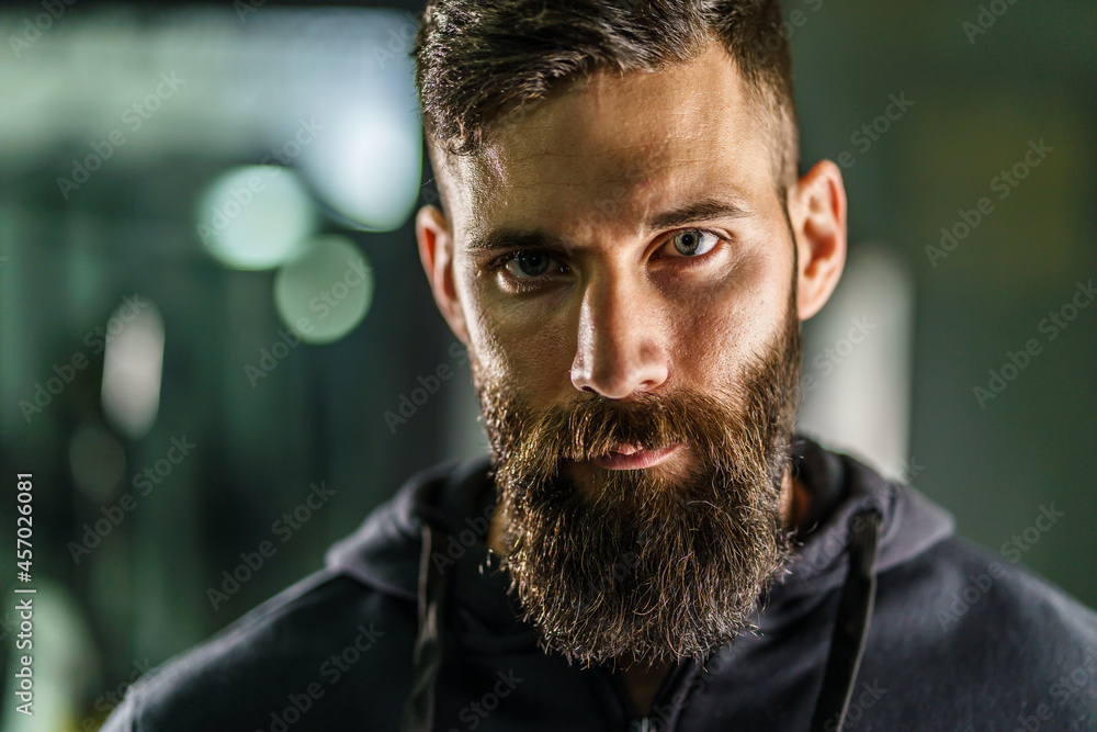 Front view close up portrait of young adult caucasian man with dark hair and beard in black shirt hoodie looking to the camera in dark - Confident modern male dramatic portrait with copy space Front view close up portrait of young adult caucasian man with dark hair and beard in black shirt hoodie looking to the camera in dark - Confident modern male dramatic portrait with copy space