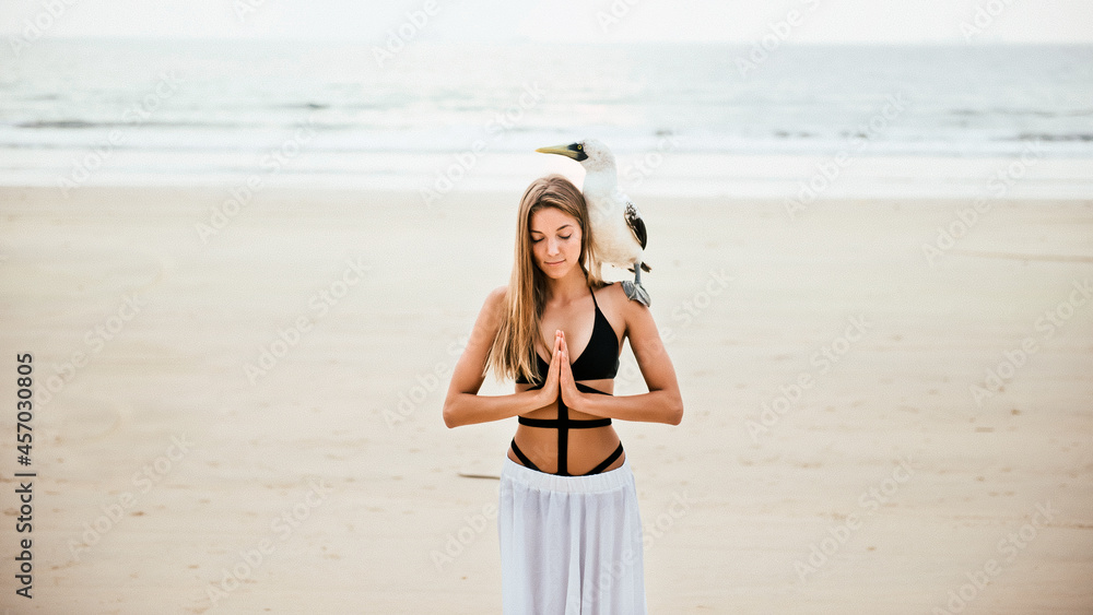 Beautiful Blonde Woman in Swimsuit Standing on Beach with Closed Eyes and Hands in Namaste Position, Large Sea Bird on Shoulde. Beautiful Blonde Woman in Swimsuit Standing on Beach with Closed Eyes and Hands in Namaste Position, Large Sea Bird on Shoulde.