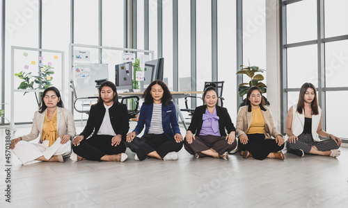 Six asian business women sitting on the floor in office with multiple glass windows meditating beside conference tables