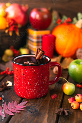 red mug with mulled wine. Cinnamon sticks stick out of the cup and a star of star anise floats. Fruits and spices are all around on a wooden table