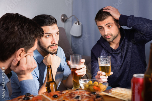 Troubled male friends talking, drinking beer with pizza at home