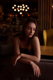  A sexy woman leans on a chair, looks down, her eyes downcast. Portrait of young woman in black dress posing in dark interior, fashion beauty photo.
