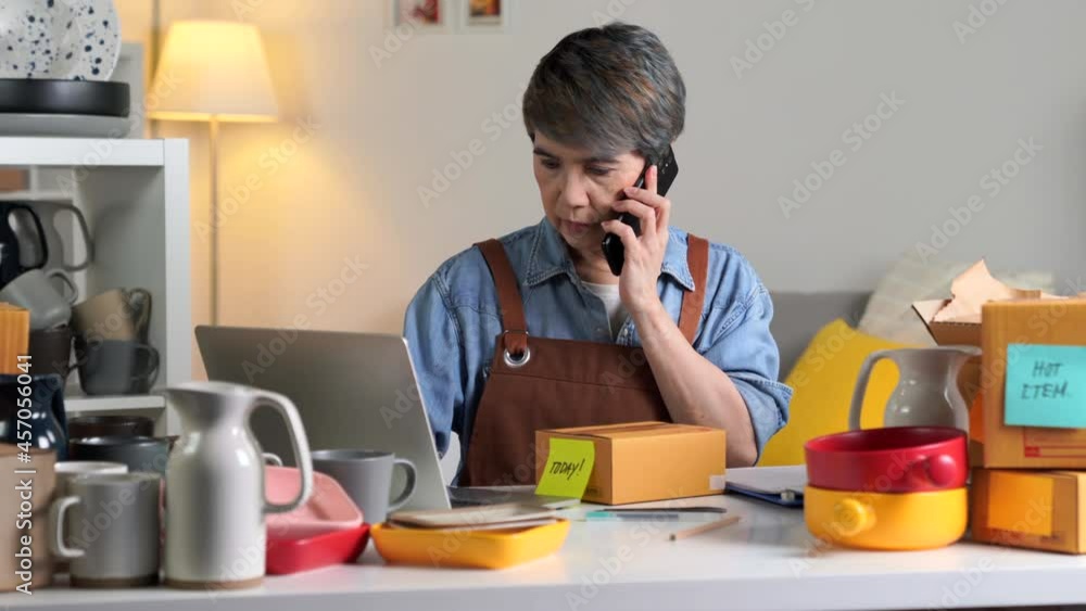 Mature Asian woman entrepreneur, Business owner on the phone with a client and checking address and delivery detail before shipping product to client. Mature Asian woman entrepreneur, Business owner on the phone with a client and checking address and delivery detail before shipping product to client.