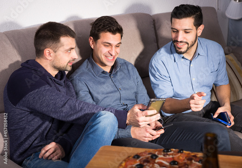 Three cheerful men using phone while enjoying beer and pizza at home