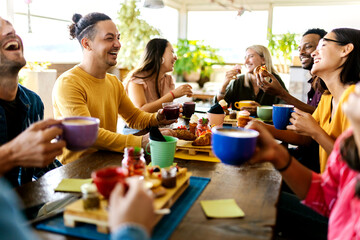 Smiling group of diverse friends having breakfast and talking at coffee bar restaurant - Happy millennial multiracial people laughing and having fun together while drinking cappuccino indoors -