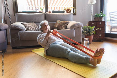 Older woman exercising with an elastic band lying on a mat on the floor of her living room at home