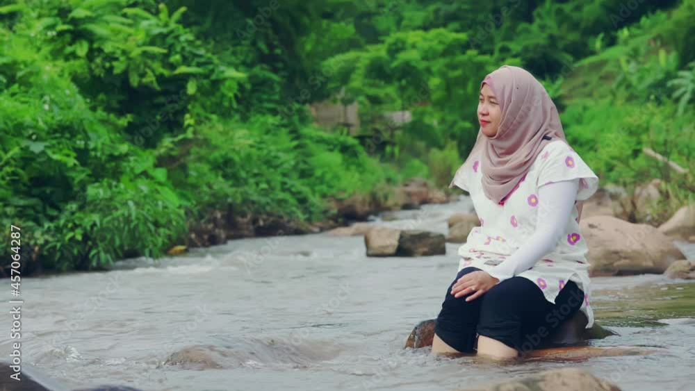beautiful Asian Muslim woman sitting on the rock with feet in a weedy pond water beautiful Asian Muslim woman sitting on the rock with feet in a weedy pond water