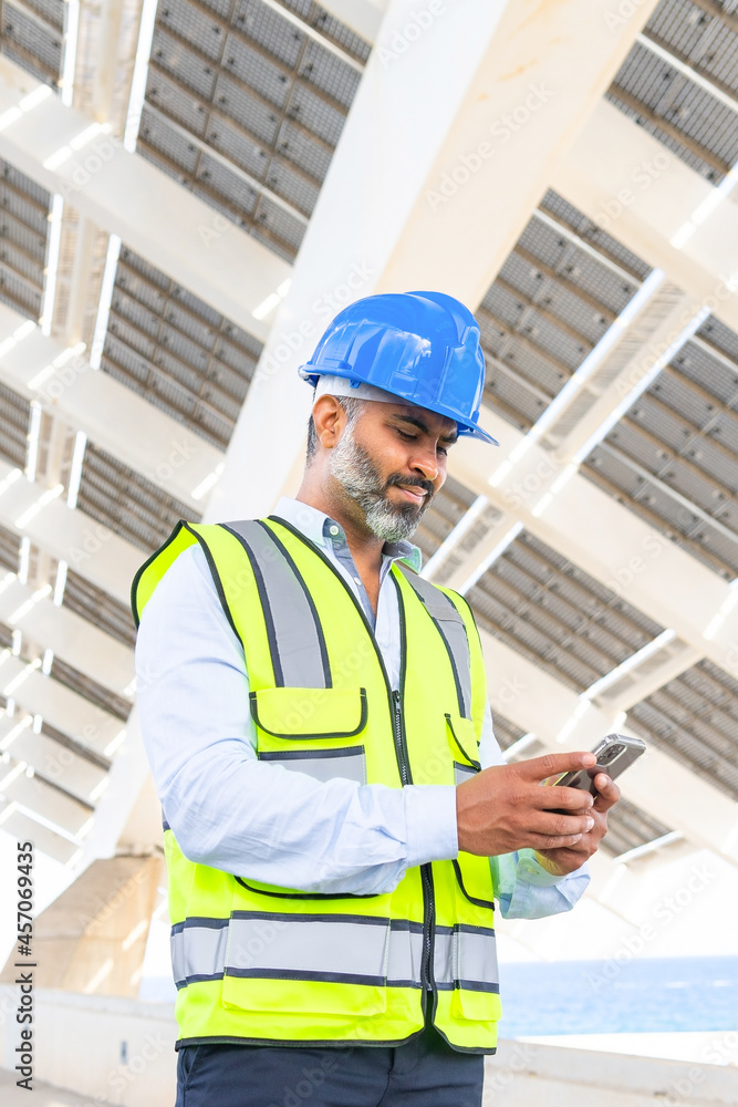 Ethnic supervisor in hardhat with smartphone against solar power station Ethnic supervisor in hardhat with smartphone against solar power station