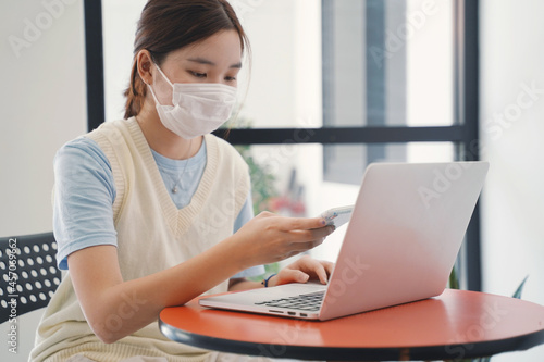 Young asian millennial woman wearing mask and using laptop and smartphone for e-learning at home, staying home and self quarantine