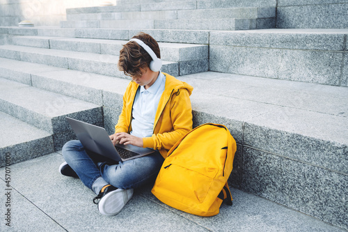 Child caucasian teen boy in yellow hoodie with headphones sitting outdoors on grey stairs using laptop. Remote or distant learning on the go. Blogging surfing. Homeschooling. Lockdown. Video call.
