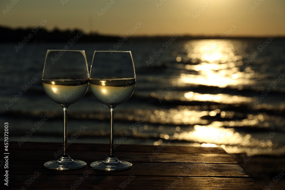 Two glasses of wine on wooden table near river at sunset. Space for text
