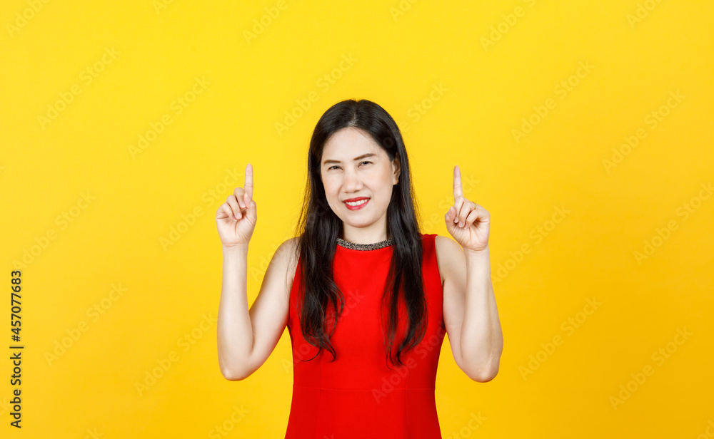 Healthy Asian woman adult wearing beautiful sleeveless dress pointing fingers to high above on bright yellow background Healthy Asian woman adult wearing beautiful sleeveless dress pointing fingers to high above on bright yellow background