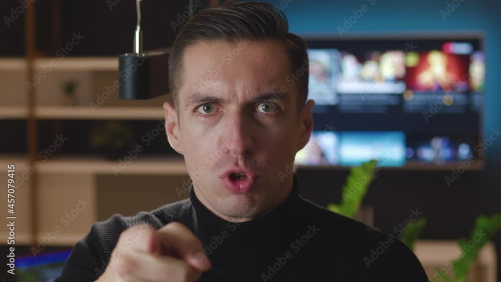 Close up angry indignant man shouting and pointing at camera at living room. Portrait of aggressive male scolding and shouting at an opponent at home in the evening Close up angry indignant man shouting and pointing at camera at living room. Portrait of aggressive male scolding and shouting at an opponent at home in the evening