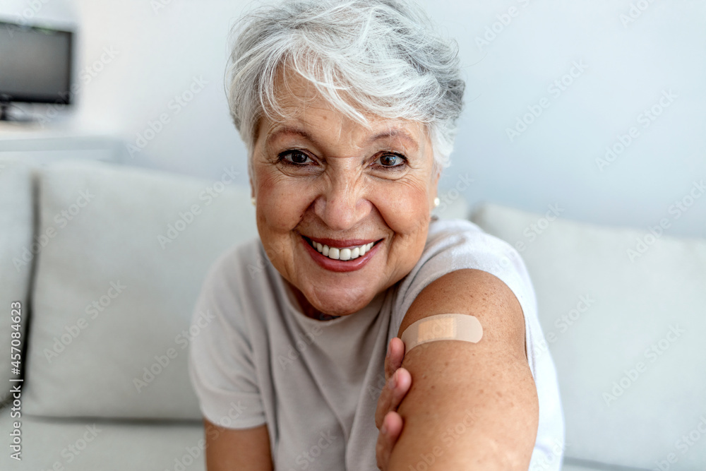 Cropped shot of a smile senior woman 70s after receiving the coronavirus covid-19 vaccine. Old aged woman posing with an adhesive COVID-19 and adhesive bandage on her upper arm. Vaccination concept. - obrazy, fototapety, plakaty 