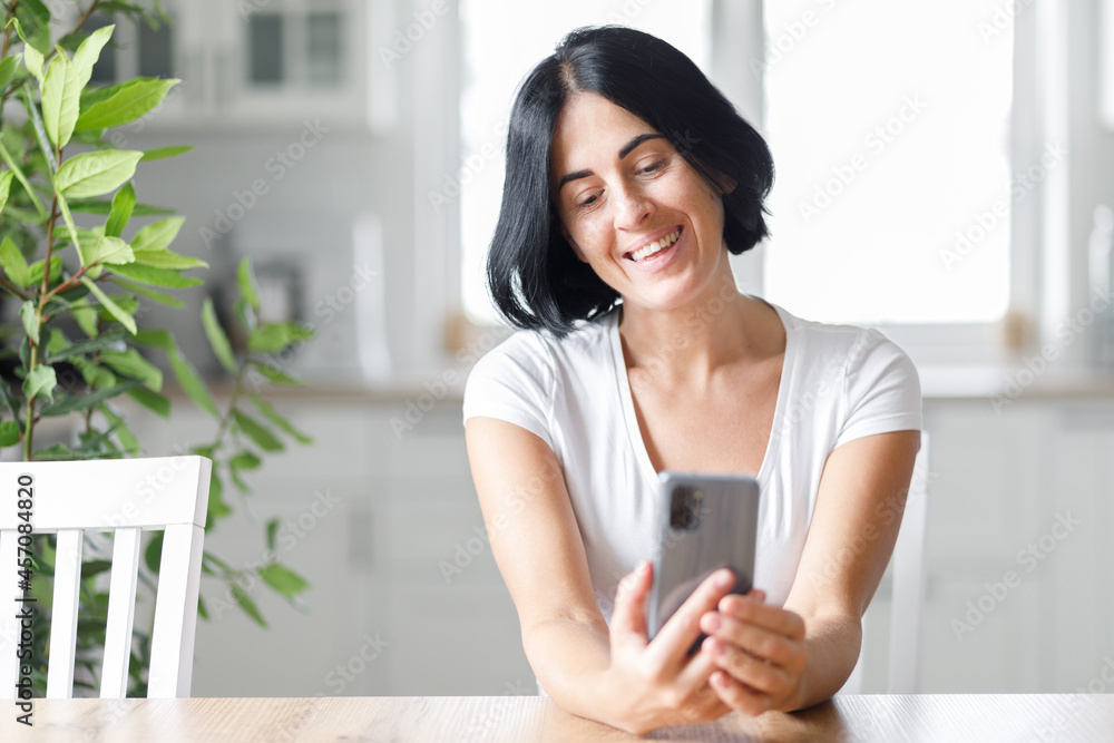 Happy woman smiling and holding smartphone in hands, young lady chatting in social networks online, watching funny videos in the internet, using mobile applications at home Happy woman smiling and holding smartphone in hands, young lady chatting in social networks online, watching funny videos in the internet, using mobile applications at home