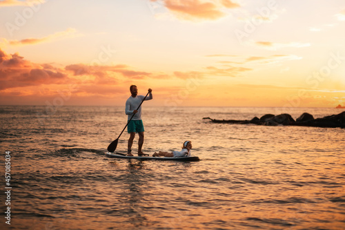 Family summer vacations. Silhouette of father and daughter surfing on a sup board. Sunset sky and sea of peach color at the background. Horizont