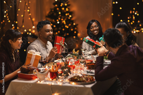 holidays  party and celebration concept - multiethnic group of happy friends having christmas dinner and giving presents at home
