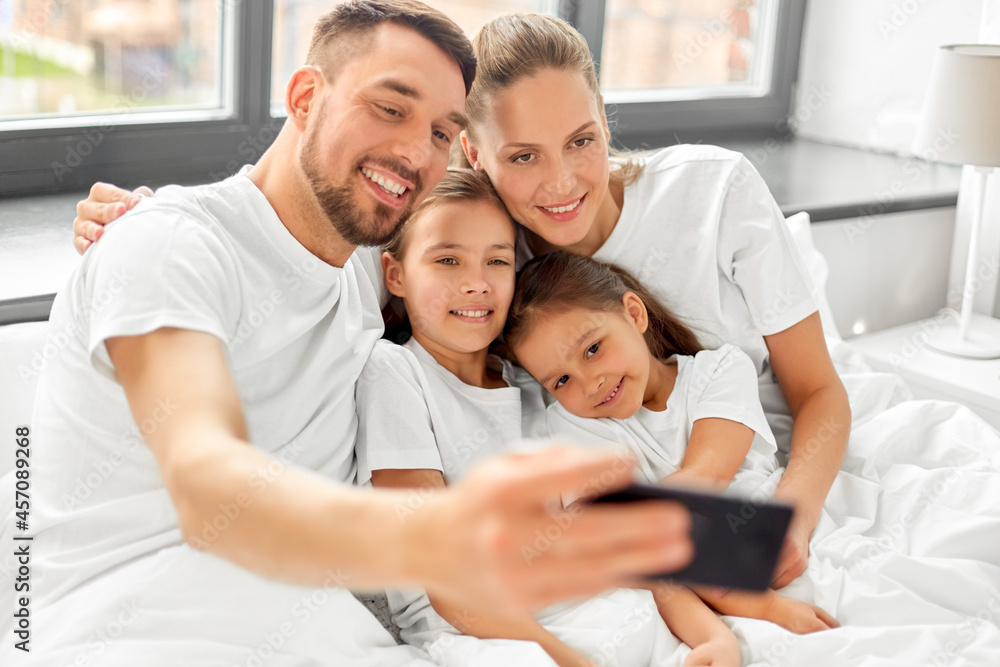 family and people concept - happy mother, father and two daughters taking selfie with smartphone in bed at home family and people concept - happy mother, father and two daughters taking selfie with smartphone in bed at home