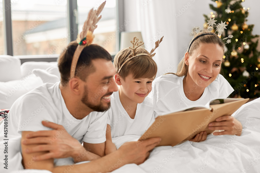 Fototapeta premium family, winter holidays and people concept - happy mother, father and little son in pajamas reading book in bed in christmas morning