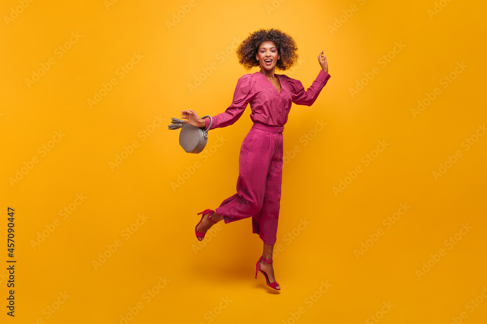 irresistible sophisticated lady jumping elegantly posing on isolated yellow background. her lush curly hair flies to sides. dressed in purple suit with handbag in her hands. irresistible sophisticated lady jumping elegantly posing on isolated yellow background. her lush curly hair flies to sides. dressed in purple suit with handbag in her hands.