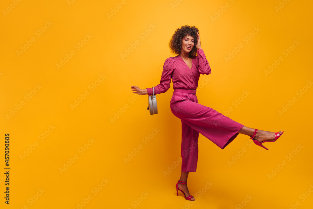  spectacular beautiful African woman raises her leg in front of her in fashionable suit and high-heeled shoes. young brown-haired woman with lush curly hairstyle with bag on her elbow in studio.