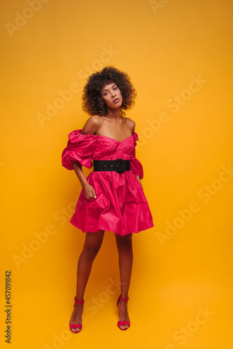 full length image of elegant dark leather model on isolated yellow background. girl in fashionable crimson dress with black belt and open shoes with heels.