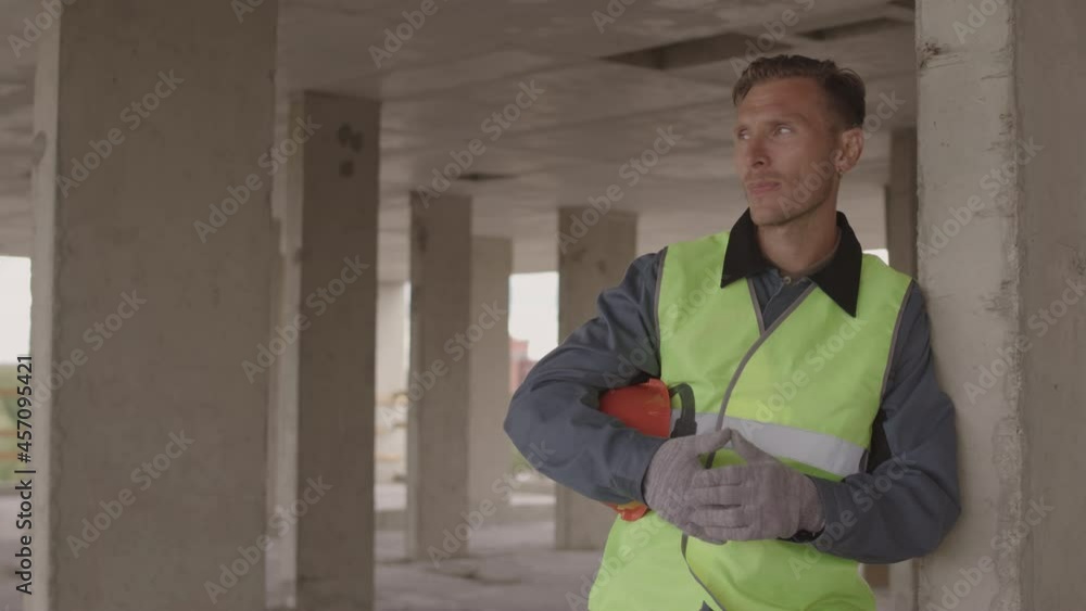 Medium of middle-aged Caucasian man wearing reflective vest, taking off hard hat, standing in building under construction, leaning on concrete pillar Medium of middle-aged Caucasian man wearing reflective vest, taking off hard hat, standing in building under construction, leaning on concrete pillar