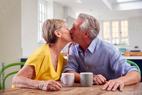 Kissing Retired Couple Sitting Around Table At Home Having Morning Coffee Together