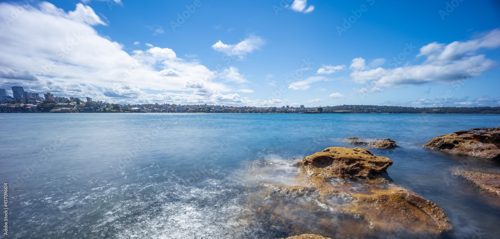 Fototapeta premium Partly cloudy Spring afternoon on Sydney Harbour with nice rocks in the foreground the soft waves crashing on the shore and the beautiful harbour foreshore as a backdrop NSW Australia