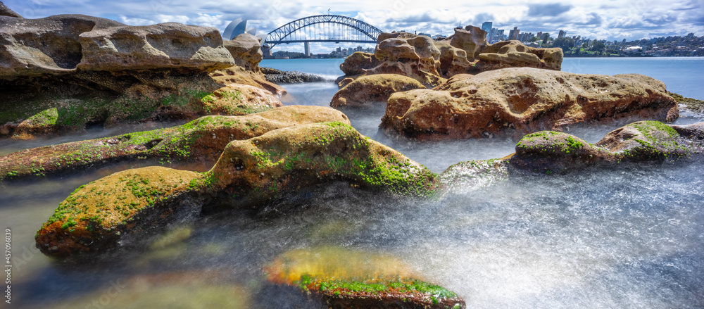 Partly cloudy Spring afternoon on Sydney Harbour with nice rocks in the foreground the soft waves crashing on the shore and the beautiful harbour foreshore as a backdrop NSW Australia