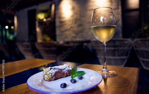 Sicilian cannoli roll with blueberries on a white ceramic plate and a glass of white wine on a restaurant table with a restaurant decor in the background.