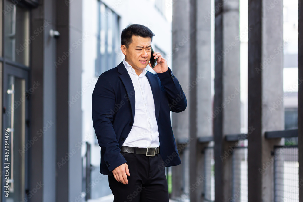 angry and serious Successful Asian businessman explains information to employees using phone, speaks near office outside angry and serious Successful Asian businessman explains information to employees using phone, speaks near office outside