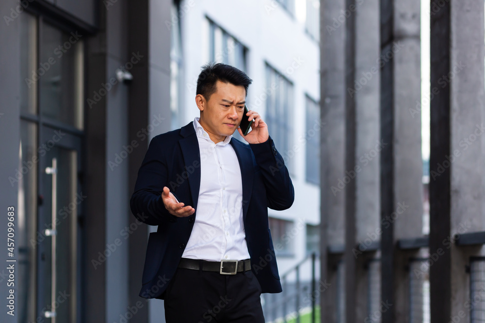 angry and serious Successful Asian businessman explains information to employees using phone, speaks near office outside angry and serious Successful Asian businessman explains information to employees using phone, speaks near office outside