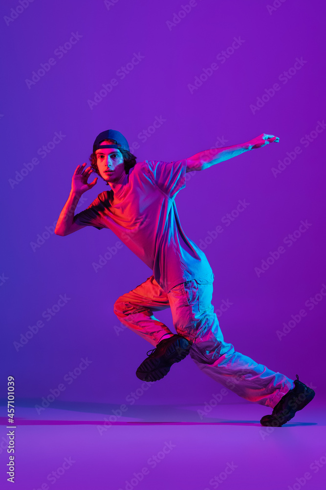 Young stylish man, break dancing, hip-hop dancer practicing solo in casual clothes isolated over purple background in neon pink light at dance hall