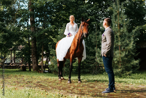 Outdoor ranch wedding. Happy young bride in white dress with bouquet sits astride horse and looks at the groom. Marriage, love and romance. Selective focus on bride