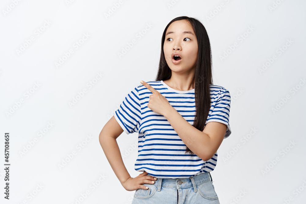 Portrait of asian girl looks surprised, say wow, pointing finger left at sale promotion, showing discount banner, standing over white background Portrait of asian girl looks surprised, say wow, pointing finger left at sale promotion, showing discount banner, standing over white background