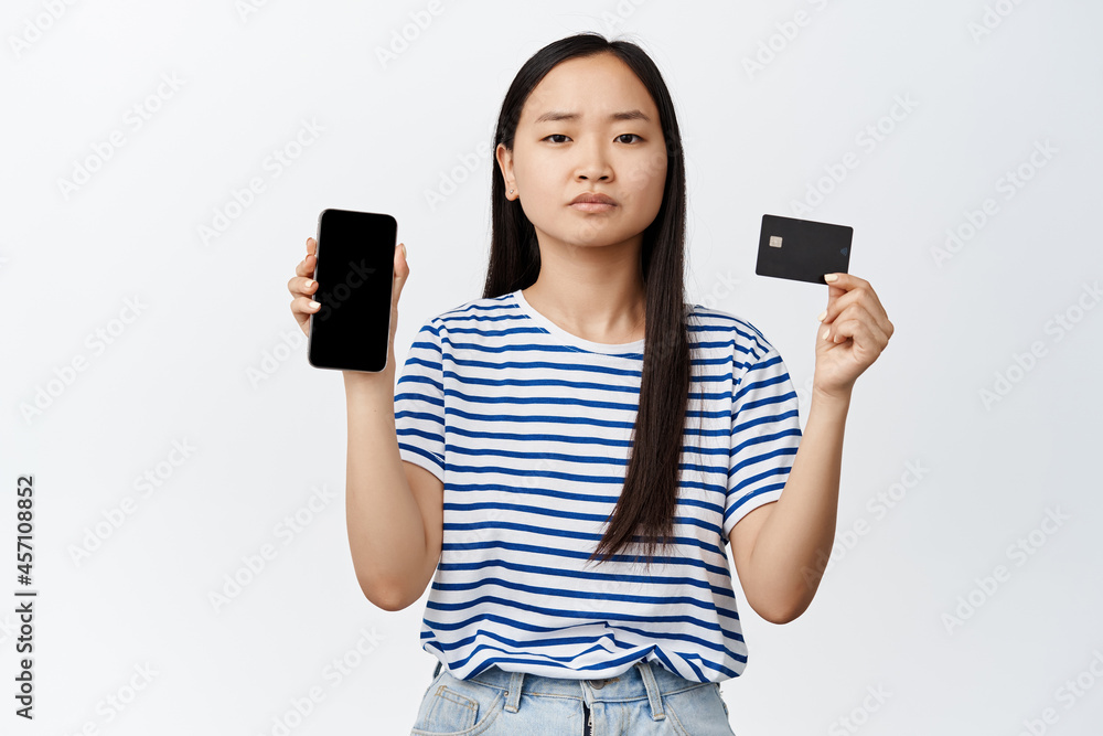Sad frowning asian girl showing mobile phone empty screen and credit card, problem with payment online, standing over white background Sad frowning asian girl showing mobile phone empty screen and credit card, problem with payment online, standing over white background
