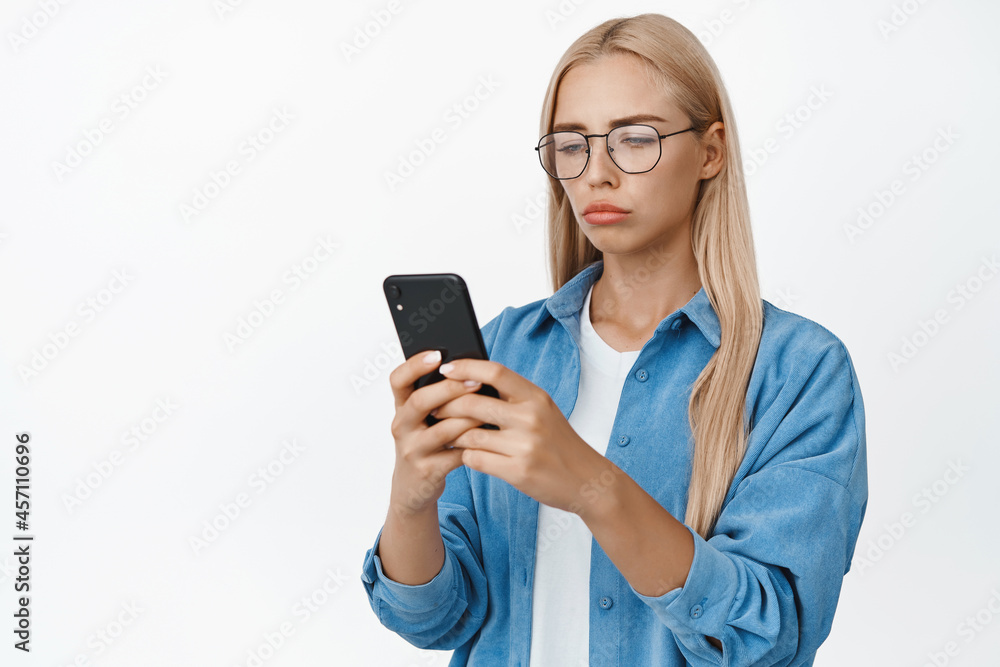 Portrait of sad and gloomy woman in glasses reading notification on mobile phone, looking at smartphone screen disappointed, white background Portrait of sad and gloomy woman in glasses reading notification on mobile phone, looking at smartphone screen disappointed, white background