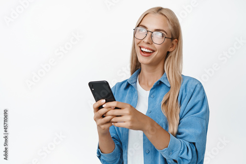 Young blond woman in glasses smiling, holding smartphone, using application with happy natural face expression, white background