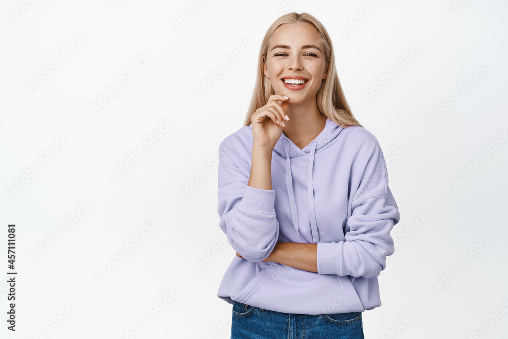 Stylish modern girl with blond long haircut, smiling at camera, looking confident, standing over white background Stylish modern girl with blond long haircut, smiling at camera, looking confident, standing over white background