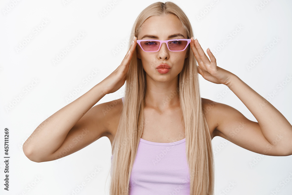 Stylish young blond woman wearing trendy sunglasses, pucker lips and looking sassy at camera, standing over white background Stylish young blond woman wearing trendy sunglasses, pucker lips and looking sassy at camera, standing over white background