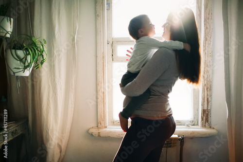 Free Download Pregnant caucasian mother with long hair with a toddler baby in her arms on the background of window in a lifestyle interior - GFXLoad