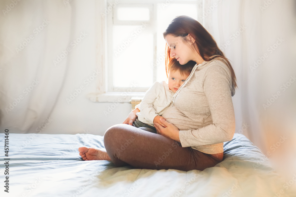 Pregnant caucasian mother with long hair embracing sad baby toddler sits on the bed by the window in a lifestyle interior, soft light and focus, lifestyle in a real bedroom Pregnant caucasian mother with long hair embracing sad baby toddler sits on the bed by the window in a lifestyle interior, soft light and focus, lifestyle in a real bedroom