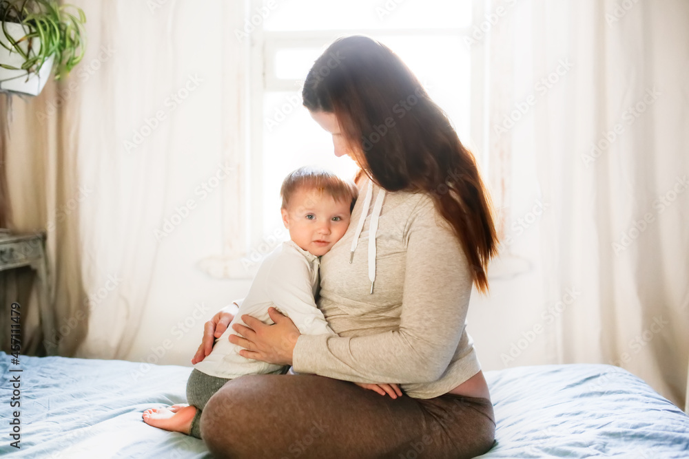 Pregnant caucasian mother with long hair embracing sad baby toddler sits on the bed by the window in a lifestyle interior, soft light and focus, lifestyle in a real bedroom Pregnant caucasian mother with long hair embracing sad baby toddler sits on the bed by the window in a lifestyle interior, soft light and focus, lifestyle in a real bedroom