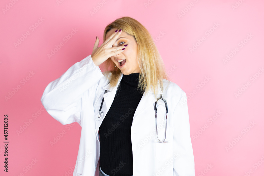 Young blonde doctor woman wearing stethoscope standing over isolated pink background peeking in shock covering face and eyes with hand, looking through fingers with embarrassed expression Young blonde doctor woman wearing stethoscope standing over isolated pink background peeking in shock covering face and eyes with hand, looking through fingers with embarrassed expression