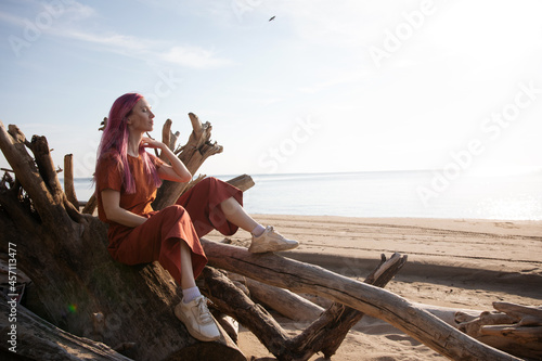 woman with pink hair sits on wooden logs on the beach and enjoys the setting sun