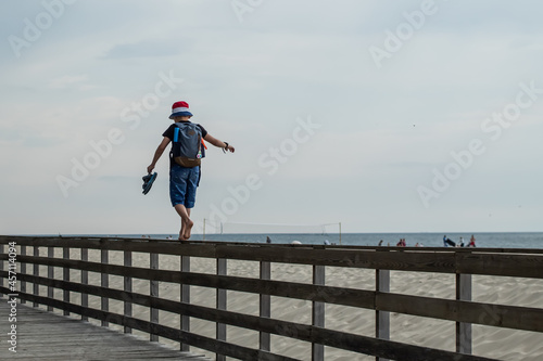 a barefoot boy in a hat and with a backpack walks away along the railing located along the beach on the sea