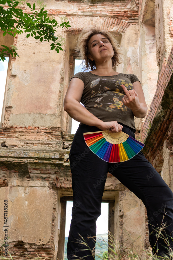 Curly haired blonde woman looking at camera covering her pubis with rainbow fan , raising middle finger in defiant attitude.