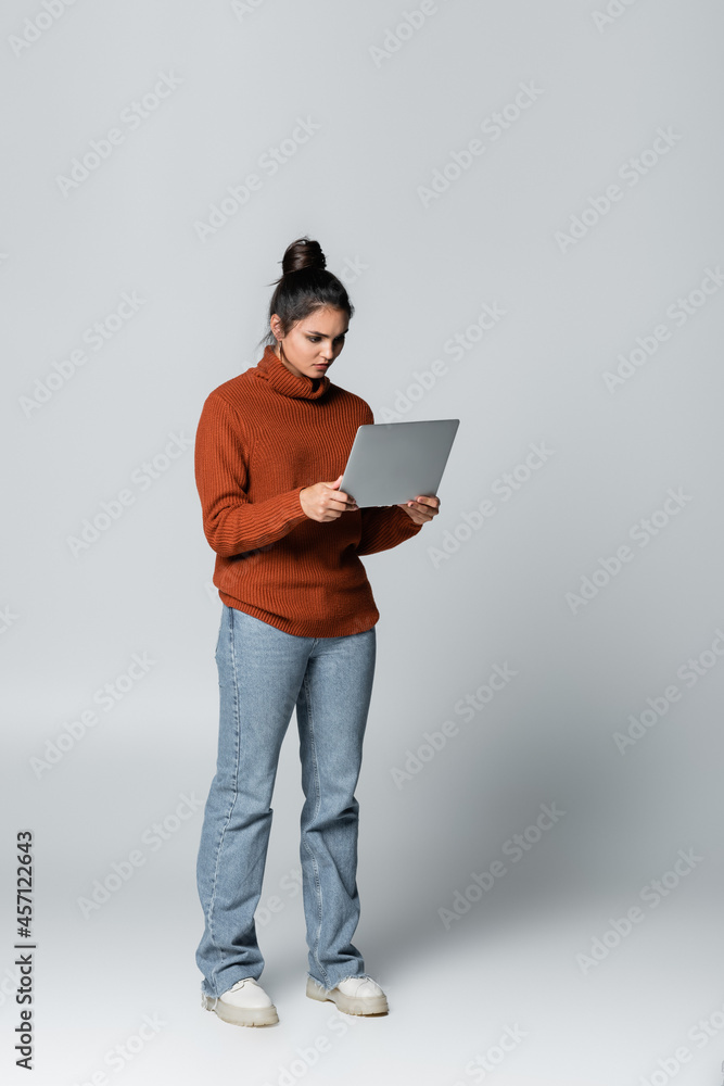 full length of young freelancer in knitted sweater and jeans using laptop on grey