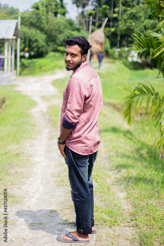 A young boy is standing in the scorching sun and looking back. his small beard and his behind blur the green nature background.
